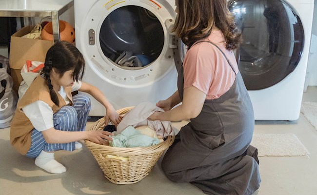 Een moeder en dochter zitten samen bij een wasmachine naast een mand met schone was.